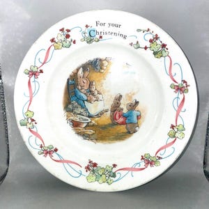 Könnte beinhalten: Ein weißer Keramikteller mit einer zentralen Illustration von Kaninchen in einem Bau. Der Text "For your Christening" ist oben aufgedruckt. Der Rand ist mit einem wiederkehrenden Muster aus rosa Schleifen, grünen Blättern und kleinen roten Blumen verziert.