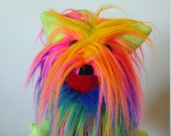May include: A bright green puppet with a long, colorful, rainbow-colored mane of fur. The puppet has a red mouth and a blue bow tie.