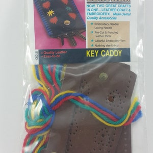 May include: A brown leather key caddy kit with embroidery floss, a needle, and pre-cut leather pieces. The kit includes instructions for making a key caddy with embroidery. The packaging says "Embroidery on Leather" and "Key Caddy".