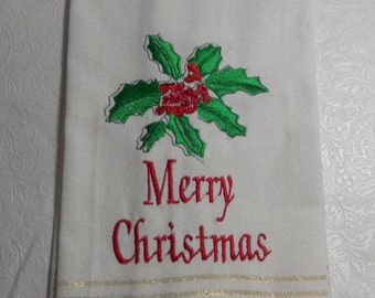 Puede incluir: Toalla de cocina blanca con rayas doradas y bordado rojo. El bordado presenta una rama de acebo con bayas rojas y el texto "Feliz Navidad".