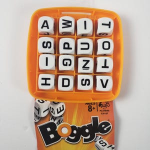 May include: An orange Boggle game set with white letter cubes. The game includes a grid base, lid, 16 letter cubes, and a sand timer. The box shows the word "Boggle" in large, black letters.