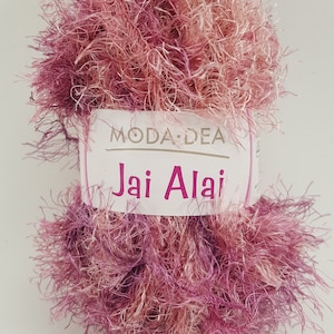 Pode incluir: Um novelo de fio felpudo rosa e roxo com a etiqueta "Moda Dea Jai Alai" nele.