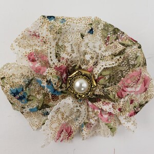 May include: A floral lace brooch with a gold-toned center and a pearl accent. The brooch features a cream-colored lace with pink, blue, and green floral patterns. The edges of the brooch are ruffled, adding texture and dimension.