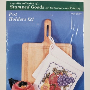 Pode incluir: Um conjunto de dois porta-panelas de madeira inacabados com um desenho de bordado de frutas. O desenho apresenta uma maçã vermelha, um pêssego amarelo e uvas roxas. A embalagem inclui o texto "Fruit 31701".