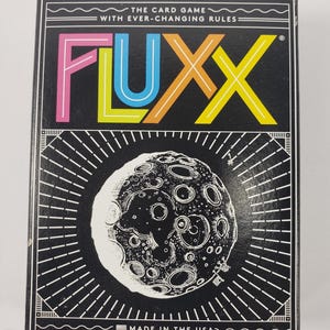 May include: A black card game box with the word "FLUX" in colorful letters. The box features a detailed illustration of the moon and the text "THE CARD GAME WITH EVER-CHANGING RULES" and "MADE IN THE USA".