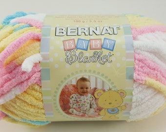 Puede incluir: Un ovillo de hilo Bernat Baby Blanket en un patrón multicolor de amarillo, rosa, blanco y azul. El hilo pesa 100 g / 3,5 oz.