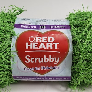 Puede incluir: Una madeja de hilo Red Heart Scrubby de color verde brillante, un tipo de hilo chenille, está lista para ser utilizada para hacer paños de cocina. La etiqueta dice "Red Heart Scrubby, ¡Ideal para paños de cocina!" e incluye información sobre el peso del hilo, el contenido de la fibra y las instrucciones de cuidado.