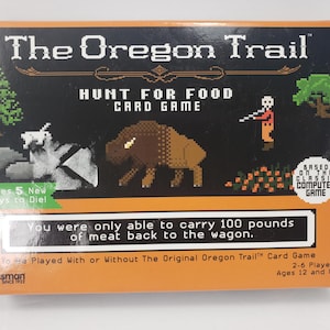 May include: The Oregon Trail Hunt for Food card game box features pixel art of a bison, a hunter, a tree, and rocks. Text reads: You were only able to carry 100 pounds of meat back to the wagon. The orange and black box indicates 2-6 players.