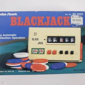 Puede incluir: Caja de juego de Blackjack Radio Shack vintage con la palabra "BLACKJACK" en letras grandes de color naranja. La caja presenta una consola de juego de color crema con pantallas numéricas y botones rojos, junto con pilas de fichas de póquer azules, rojas y blancas.