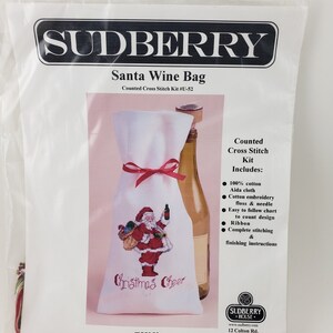 May include: A counted cross stitch kit for a Santa wine bag. The kit includes white Aida cloth, red embroidery floss, a needle, and instructions. The design features a Santa Claus holding a bottle of wine and a bag of gifts. The text "Christmas Cheer" is stitched below the image.