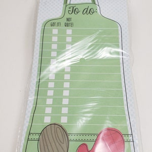 May include: A green magnetic notepad with a kitchen apron design. The notepad has a checklist with the words "To do: Got it! Not Quite!" and includes a red oven mitt and a wooden spoon.