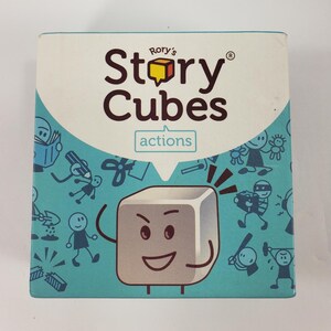May include: A square box for Rory's Story Cubes, with the text "Story Cubes" and "actions" printed on the front. The box has a white background with a teal border and cartoon illustrations of stick figures and objects.
