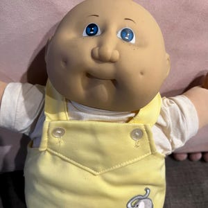 May include: A vintage baby doll with a bald head, blue eyes, and rosy cheeks. The doll is wearing a yellow overall with a white shirt underneath. An embroidered elephant is on the front of the overall. The doll's arms are outstretched.