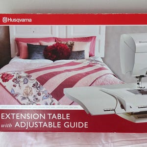 May include: A rectangular red and white product box for a Husqvarna Viking extension table with adjustable guide. The box features a graphic of a white sewing machine attached to the extension table, set against a bedroom scene with a pink quilt.