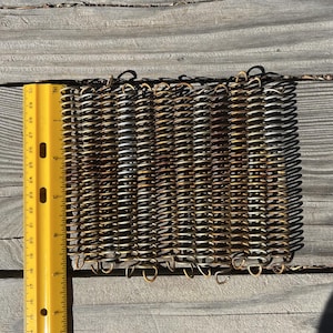 May include: A close-up of a woven metal mesh mat with a ruler for scale. The mat is made of small, interconnected metal rings, creating a grid-like pattern. The metal is a mix of silver and gold tones, with some areas showing signs of rust.