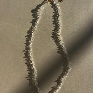 Könnte beinhalten: Eine zarte Halskette aus kleinen, klaren, facettierten Perlen. Die Halskette hat einen goldfarbenen Verschluss und eine Kette. Die Perlen fangen das Licht ein und erzeugen einen funkelnden Effekt. Die Halskette wird vor einem neutralen Hintergrund präsentiert.