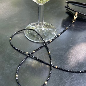Puede incluir: Una cadena de gafas de cuentas negras con detalles dorados. La cadena está enroscada alrededor de una copa de vino.
