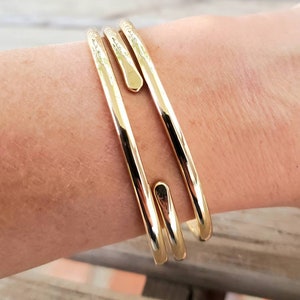 May include: Three gold-toned bangle bracelets stacked on a person's wrist. The bracelets are smooth and have a rounded shape.