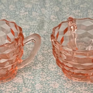 May include: A pink glass creamer and sugar bowl set. The creamer has a handle and a spout. Both pieces have a diamond pattern. The set is on a light blue floral patterned surface.