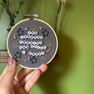 May include: A gray embroidery hoop with the text "THE HORRORS PERSIST AND STILL I CRAFT" in white beads. The design includes embroidered stars, rainbows, and other small embellishments in various colors. The hoop is held by a hand.