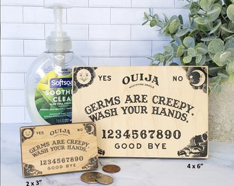 Könnte beinhalten: Zwei Holzschildchen mit dem Text "Ouija Mystifying Oracle" und "Germs are creepy. Wash your hands." Die Schilder sind mit einer Sonne, einem Mond und Sternen verziert. Das größere Schild ist 4 Zoll mal 6 Zoll groß und das kleinere Schild ist 2 Zoll mal 3 Zoll groß.