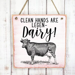 May include: A 7x7 inch wooden sign with a pink background and white text that reads "Clean Hands Are Legen-Dairy!" with a black and white illustration of a cow. The sign is hanging from twine.