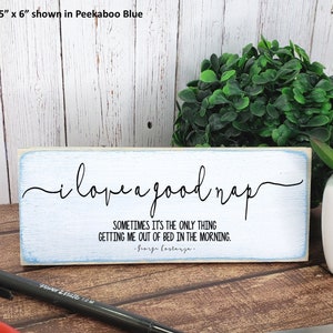 May include: A white wooden sign with blue trim reads "i love a good nap" in a cursive font. The sign also includes the quote "Sometimes it's the only thing getting me out of bed in the morning." attributed to "George Costanza."