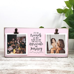 Pode incluir: Uma moldura dupla rosa com uma moldura de madeira branca. A moldura tem duas aberturas de 10x10 cm com clipes para segurar fotos. A moldura tem uma citação que diz "São os amigos que encontramos ao longo do caminho que nos ajudam a apreciar a jornada".