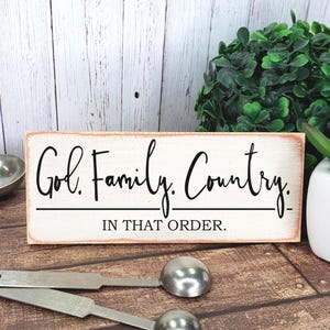 May include: A rectangular wooden sign with the words "God. Family. Country. IN THAT ORDER." in black script. The sign is 2.25 inches high and 6 inches wide. The background is white with a distressed look, and the edges are stained.