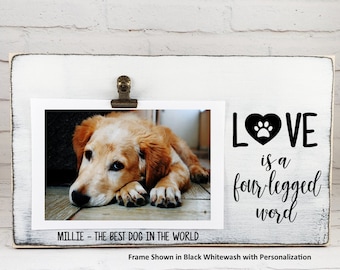 May include: White wooden picture frame with a black and whitewash finish. The frame has a clip to hold a photo. The photo shows a golden retriever puppy. The text on the frame reads "LOVE is a four legged word".