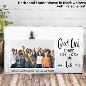 May include: Whitewashed wooden picture frame with a black clip holding a photo of a group of people. The frame has a black inscription that reads "Good Luck Finding A Better Class Than -US- ".