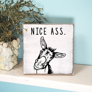 May include: A white wooden sign with black text and a donkey illustration. The sign reads "NICE ASS." The sign is 7 inches by 7 inches and is shown in black whitewash.