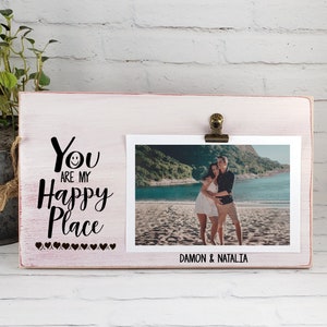 Puede incluir: Un marco de fotos de madera blanca con un clip en la parte superior. El marco tiene una inscripción negra que dice "You are my Happy Place" con una carita sonriente y corazones. Una foto de una pareja en una playa se muestra en el marco. Los nombres "Damon & Natalia" están escritos debajo de la foto.