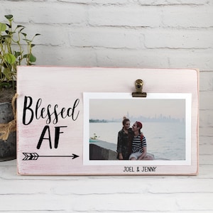 May include: A pink wooden picture frame with a white clip holding a photo of a couple. The frame has the text "Blessed AF" with an arrow pointing to the right. The photo is of a couple standing on a pier with a city skyline in the background. The text "Joel & Jenny" is printed below the photo.
