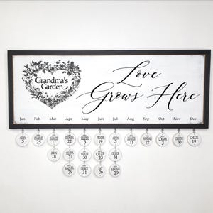May include: A black framed calendar with the text "Love Grows Here" and "Grandma's Garden". The calendar is 8 x 22 inches and has personalized disks with names and dates. The design includes a floral heart.