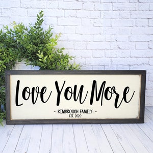 May include: A rectangular framed sign with the words "Love You More" in black script. Below the text is "~ KIMBROUGH FAMILY ~ EST. 2020". The sign is 6x16 inches and shown in Peppered Cream. A green wreath is in the background.