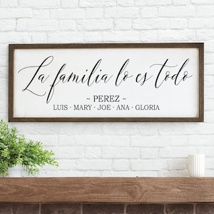 May include: A rectangular wooden sign with the phrase "La familia lo es todo" in elegant black script. Below, the family name "PEREZ" and individual names are listed. The sign is 10 x 26 inches and is displayed against a white brick wall.