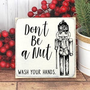 May include: A white wooden sign with black text and a black and white illustration of a nutcracker. The sign reads "Don't Be a Nut Wash Your Hands."