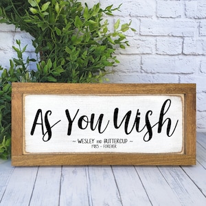 May include: A framed wooden sign with white background and black text that reads "As You Wish ~ WESLEY AND BUTTERCUP ~ 1985 - FOREVER". The sign is 3x8 inches and is shown in American Boho style.