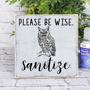 May include: A white wooden sign with black text that reads "PLEASE BE WISE. Sanitize."  An illustration of an owl is above the text. The sign is 5x5 inches and is shown in a foggy night setting.