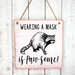 May include: A 7x7 inch wooden sign with a pink background and white painted text that reads "WEARING A MASK is Paw-Some!"  A black and white illustration of a raccoon wearing a mask is in the center of the sign.