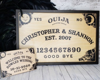 May include: A wooden Ouija board with black lettering and a dark frame. The board includes the words "Christopher & Shannon Est. 2007" and numbers. A smaller sign reads "Welcome to the Rowles Wedding 10-31-2020."