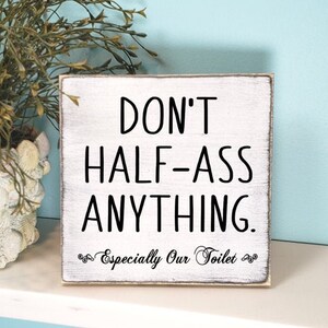 May include: A whitewashed wooden sign with black text that reads "DON'T HALF-ASS ANYTHING. Especially Our Toilet." The sign is 7 inches by 7 inches and is shown in black whitewash.