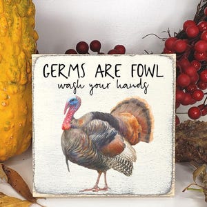 May include: A square sign with the text "GERMS ARE FOWL wash your hands" and a watercolor illustration of a turkey. The sign is 5x5 inches and has a distressed, cream-colored background.