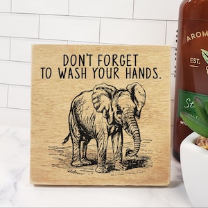 May include: A wooden sign with a brown background and black text that reads "Don't forget to wash your hands."  The sign features a black and white illustration of an elephant. The sign is 5 inches by 5 inches.
