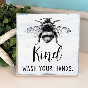 May include: A 5x5 inch wooden sign with a blue background and a black and white illustration of a bumblebee. The sign reads "Kind. Wash Your Hands."