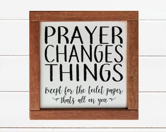 May include: A white wooden sign with a brown frame. The sign says "PRAYER CHANGES THINGS Except for the toilet paper That's all on you".