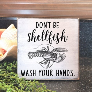 May include: A whitewashed wooden sign with black text that reads "Don't be shellfish. Wash your hands."  A black and white illustration of a lobster is featured on the sign.