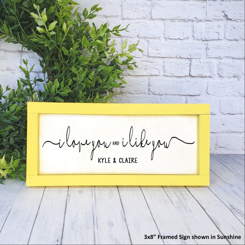 May include: A yellow framed sign with the text "i love you and i like you" with the names "KYLE & CLAIRE" underneath. The sign is 3x8 inches and is shown against a white brick wall.