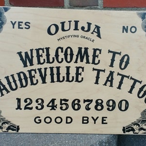 Pu&ograve; includere: Una tavola Ouija in legno con scritte e illustrazioni nere. La tavola dice "Ouija Mystifying Oracle" e "Welcome to Vaudeville Tattoo". Presenta i numeri 0-9, "Yes", "No" e "Good Bye".
